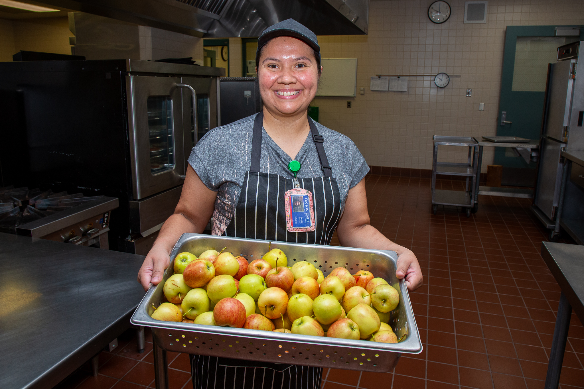 A woman smiles as she holds up a bin of apples in the cafeteria